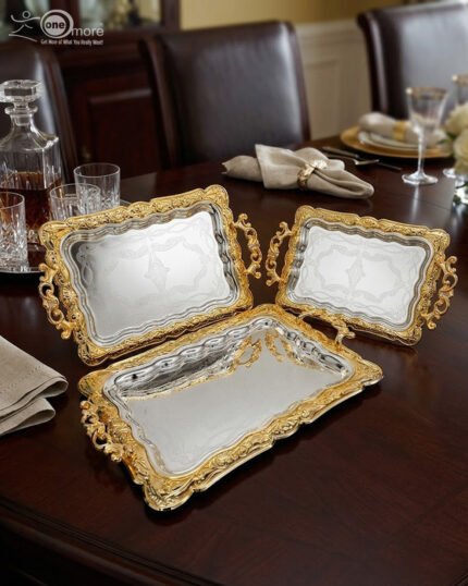 A three-piece set of rectangular stainless steel serving trays featuring polished silver mirror bases with etched ribbon designs and ornate gold-embossed scalloped borders, displayed on a dark wooden dining table.