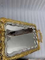 A three-piece set of rectangular stainless steel serving trays featuring polished silver mirror bases with etched ribbon designs and ornate gold-embossed scalloped borders, displayed on a dark wooden dining table.