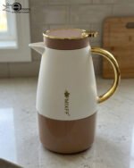 Three elegant 1.5-liter stainless steel thermal flasks in rose gold, slate grey, and charcoal blue bases with white bodies and gold-plated handles, displayed on a marble kitchen countertop.