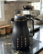 Two elegant 1-liter thermal carafes in matte black and slate blue with gold decorative patterns, displayed on a kitchen counter next to a coffee machine.