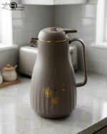 Two 1-liter insulated thermal flasks in Earth Grey and Creamy Beige, featuring golden dandelion designs and ergonomic handles, displayed on a kitchen countertop.