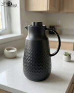 A 1-liter insulated thermal flask with a 3D diamond-patterned matte body, featuring a curved handle and a spout, shown in black and beige color options on a kitchen countertop.