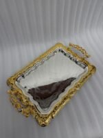 A three-piece set of rectangular stainless steel serving trays featuring polished silver mirror bases with etched ribbon designs and ornate gold-embossed scalloped borders, displayed on a dark wooden dining table.