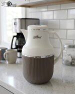 Three 1-liter modern vacuum flasks with cream-colored tops and ribbed bases in brown, grey, and beige, displayed on a white marble kitchen countertop.