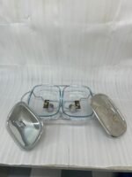 A Paramount Series double stainless steel food warmer on a dining table, featuring two 1.5L glass dishes with lids being lifted to reveal steaming food, supported by a chrome frame with two lit buffet burners underneath.