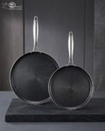 A professional-grade laser-etched non-stick stainless steel crepe pan with a textured honeycomb surface and ergonomic silver handle, shown on a kitchen counter and an induction cooktop.