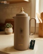 Three 1-liter DayDays insulated flasks in white, taupe, and grey, featuring gold floral designs and decorative gold lids on a kitchen counter.