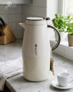 Three 2-liter luxury stainless steel thermal flasks in black, cream, and grey finishes with metallic handles, arranged on a white marble kitchen counter.