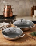 High-angle shot of two Uakeen stainless steel non-stick woks in 30cm and 32cm sizes on a wooden countertop, showing the laser-etched hexagonal interior and tempered glass lids.