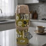A 1.9-liter olive green Day Days vacuum insulated coffee airpot with a brown pump-action lid and handle, featuring a decorative coffee bean pattern, displayed on a kitchen counter next to a coffee cup.