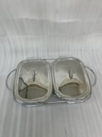 A Paramount Series double stainless steel food warmer on a dining table, featuring two 1.5L glass dishes with lids being lifted to reveal steaming food, supported by a chrome frame with two lit buffet burners underneath.