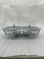 A Paramount Series double stainless steel food warmer on a dining table, featuring two 1.5L glass dishes with lids being lifted to reveal steaming food, supported by a chrome frame with two lit buffet burners underneath.