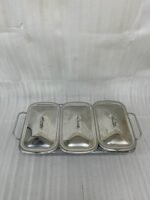 A professional Paramount Series 3-in-1 stainless steel food warmer with three 1.5L glass containers and polished metal lids, displayed on a kitchen counter and a dining table with various dishes.