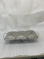 A professional Paramount Series 3-in-1 stainless steel food warmer with three 1.5L glass containers and polished metal lids, displayed on a kitchen counter and a dining table with various dishes.