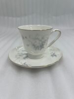A person holding a white bone china teacup filled with milk tea, featuring a grey floral design and gold rim, resting on a matching saucer with a silver spoon on a wooden table.