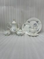 A luxury white super bone china tea set with grey floral patterns and gold rims on a wooden table, featuring a teapot, teacup, plates, and a steaming cup of tea against a snowy window BG.
