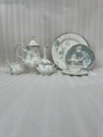 A premium super bone china tea set featuring a classic teapot, sugar bowl, creamer, teacup with saucer, and matching plates, all adorned with an elegant light blue and grey floral pattern on a wooden table.