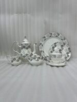 A premium bone china tea and coffee set with a grey floral pattern and silver rims on a dark wooden table. The collection includes a teapot, creamer pitcher, sugar bowl, plates, and a steaming cup of black coffee on a saucer with a silver spoon, set against a bright window showing a lush garden background