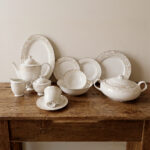 A comprehensive 61-piece super bone china dinner set arranged for display against a neutral textured background. The white porcelain tableware features a delicate cream and gold floral pattern on the rims. The collection includes dinner plates, side plates, soup bowls, a large oval platter, a lidded serving tureen, a teapot with matching sugar bowl and creamer, and teacups with saucers
