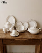 A comprehensive 61-piece super bone china dinner set arranged for display against a neutral textured background. The white porcelain tableware features a delicate cream and gold floral pattern on the rims. The collection includes dinner plates, side plates, soup bowls, a large oval platter, a lidded serving tureen, a teapot with matching sugar bowl and creamer, and teacups with saucers