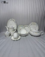 A detailed product photograph of a 61-piece 'super bone' china dinner set by One More, arranged on a white marble countertop against a neutral background. The comprehensive set, with service for 8 people, includes a teapot, creamer, sugar bowl with lid, tea cup with saucer, a large lidded casserole dish, numerous plates (dinner, salad, soup) and bowls stacked, and an oval serving platter. Each piece features a matching, delicate blue and grey floral motif with a refined gold rim, presented under elegant natural lighting. The One More logo is in the upper left