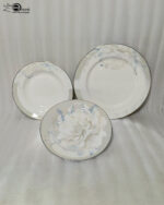 A close-up photograph of a massive, multi-tiered stack of sophisticated grey and white dinner plates featuring a detailed peony floral design with blue accents and silver rims. The plates are neatly arranged on a grey linen runner on a dining table, next to a crystal wine glass and another set of plates. The brand "One More" logo is visible in the background