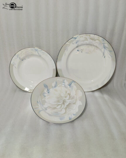 A close-up photograph of a massive, multi-tiered stack of sophisticated grey and white dinner plates featuring a detailed peony floral design with blue accents and silver rims. The plates are neatly arranged on a grey linen runner on a dining table, next to a crystal wine glass and another set of plates. The brand "One More" logo is visible in the background