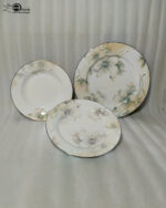 A beautifully stacked 18-piece premium Super Bone China plate set featuring an elegant floral pattern and fine trim, arranged on a beige linen table runner alongside crystal wine glasses and a rustic flower vase.