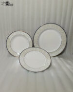 A professionally styled stack of the OneMore 18-piece super bone china dinnerware set, showcasing six quarter plates on top, six deep plates in the middle, and six large rice plates at the bottom, featuring an elegant light-green patterned border on a silver table runner