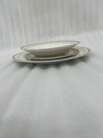 A stacked 18-piece white Super Bone China dinner plate set with elegant gold borders, arranged neatly on a polished wooden dining table alongside a wine glass and silverware under warm chandelier lighting.