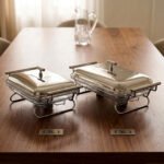 Two Paramount Series stainless steel food warmers in 2L and 3L sizes on a wooden table, featuring glass basins and polished metal lids.
