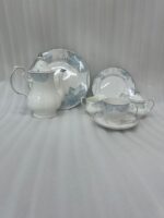 A 24-piece luxury porcelain tea set featuring a delicate blue floral pattern and silver trim, including a teapot, cups, saucers, plates, sugar bowl, and milk jug on a linen runner.