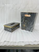 A close-up photograph of a hand pulling a tissue from a luxury black marble and gold-veined tissue box cover with gold trim, next to a matching small open-top waste bin with a warm terracotta interior and cutout handles, all on a wooden surface in a contemporary bathroom setting.