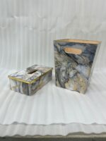 A hand dropping a tissue into a modern marble-patterned dustbin with gold trim, sitting next to a matching tissue box cover on a wooden counter.