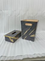 A hand with a gold ring is seen dropping a crumpled tissue into a sophisticated dark marble waste bin with prominent gold veining and trim. In the foreground is a matching tissue box cover on a wooden desk. A stacked notebook and leather portfolio are also visible.