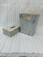 A close-up view of a luxury faux-marble waste bin with intricate gold veining and polished gold trim, positioned next to a matching marble tissue box holder on a wooden desk. A hand is seen depositing a tissue into the bin, which features a peach-pink interior lining.