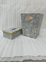 A close-up view of a hand reaching to pull a tissue from a grey and gold-veined faux-marble tissue box cover with polished gold trim, next to a matching compact grey marble waste bin with handle cutouts and a peach-colored interior, sitting on a wooden desk.