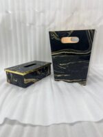 Luxury black and gold marble pattern dustbin and matching tissue box holder on a wooden vanity next to a marble bathroom sink.