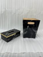 A matching executive desk accessory set featuring a black marble pattern wastebasket and a rectangular tissue box cover with elegant gold trim, placed on a dark wooden office desk.