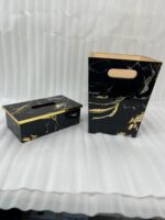 A hand pulling a tissue from a luxury black and gold marble tissue box cover next to a matching wastebasket on a table.