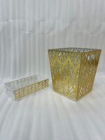An elegant gold and clear acrylic dust bin with a laser-cut leaf pattern on a wooden table, featuring a matching tissue box holder and a hand dropping a crumpled tissue inside.