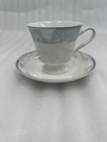 Elegant super bone china tea cup and saucer with a light blue floral pattern and gold-trimmed handle, placed on a wooden table next to a silver spoon