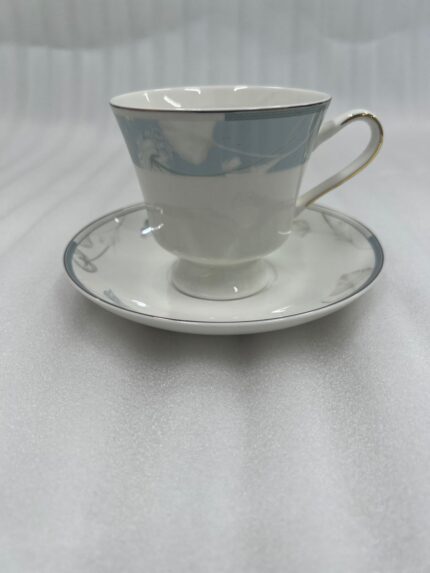 Elegant super bone china tea cup and saucer with a light blue floral pattern and gold-trimmed handle, placed on a wooden table next to a silver spoon