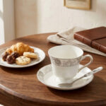 Elegant white Super Bone China teacup and saucer featuring a grey floral pattern and gold rim, placed on a wooden table next to a silver spoon and lavender vase