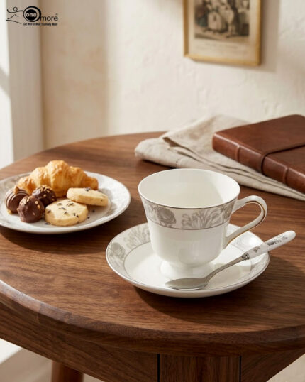 Elegant white Super Bone China teacup and saucer featuring a grey floral pattern and gold rim, placed on a wooden table next to a silver spoon and lavender vase
