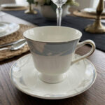 Elegant super bone china tea cup and saucer with a light blue floral pattern and gold-trimmed handle, placed on a wooden table next to a silver spoon