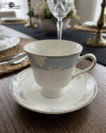Elegant super bone china tea cup and saucer with a light blue floral pattern and gold-trimmed handle, placed on a wooden table next to a silver spoon