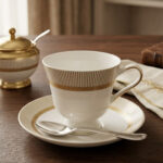 A premium white Super Bone China tea cup and saucer set featuring gold vertical stripes and a gold laurel wreath pattern on a dark wooden table