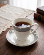Elegant white Super Bone China tea cup and saucer with a grey floral pattern and gold rim, filled with tea and garnished with star anise and a jasmine flower on a wooden table