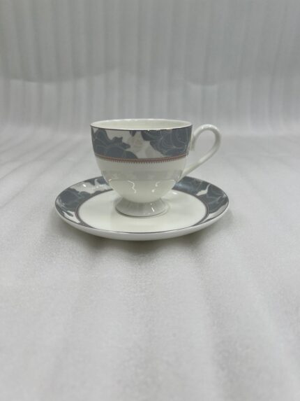 A high-angle shot of a premium super bone china cup and saucer filled with milk tea, featuring a grey floral pattern and silver spoon, set on a wooden table in a cozy living room with green sofas and a fireplace.